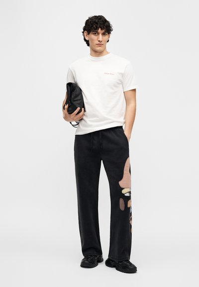 Άσπρο βαμβακερό μπλουζάκι με κόκκινο λογότυπο· μαύρο παντελόνι φόρμας με πολύχρωμα αφηρημένα μπαλώματα· το μοντέλο κρατάει μια μαύρη τσάντα· φορεμένο με μαύρα παπούτσια.