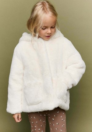Jeune fille portant une veste blanche à capuche duveteuse et des leggings floraux marron, regardant vers le bas avec une main dans la poche de la veste.