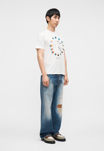 Λευκό βαμβακερό T-shirt με κυκλικό πολύχρωμο σχέδιο και κείμενο. Φορεμένο με ταλαιπωρημένα μπλε τζιν παντελόνια με επιδιορθωτικές ραφές και καφέ παπούτσια.