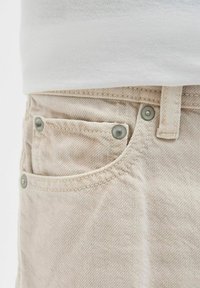 Pantalons en denim beige avec une poche avant dotée de boutons en métal argenté. Le tissu présente un motif texturé subtil et des détails de couture visibles.