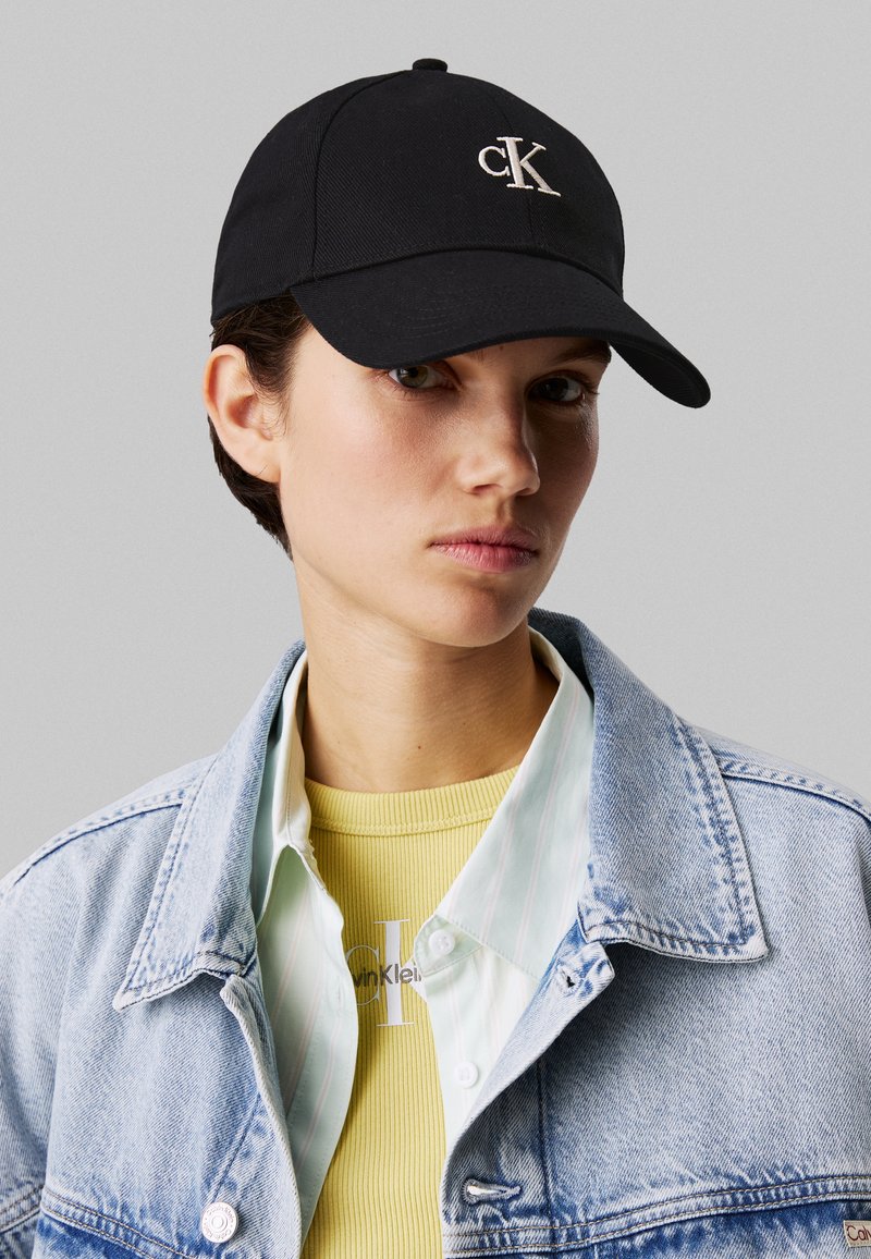 Black cotton cap featuring an embroidered "ck" logo. Model wears a light denim jacket over a yellow ribbed shirt and striped shirt.