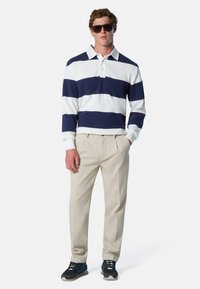 Mann mit dunklen Sonnenbrillen, dunkelblauem und weiß gestreiften Rugby-Shirt, beigen Hosen und schwarzen Sneakern, der mit einer Hand in der Tasche steht.