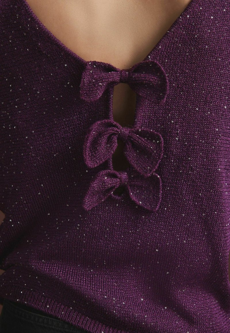Gros plan d'un vêtement tricoté violet avec de petites paillettes et trois nœuds en tissu noués à un décolleté profond en V, laissant apparaître la peau en dessous.