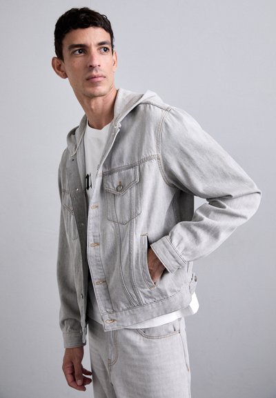 Lys grå denimjakke med hætte, der har to brystlommer, beige syninger og en afslappet pasform. Bæres over en hvid grafisk t-shirt.