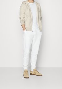 Man i beige hoodie, vit t-shirt, vita joggers och beige sneakers som står mot en vit bakgrund.