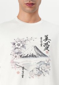 T-shirt en coton blanc avec un design graphique du mont Fuji entouré de fleurs et de texte japonais dans des tons noirs et roses.