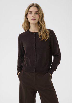 Femme aux longs cheveux blonds portant un cardigan boutonné marron foncé et un pantalon assorti, debout les mains dans les poches devant un fond uni.