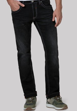 Man met zwarte jeans met contrasterende stiksels en groene sportschoenen, staand met één hand in de zak tegen een grijze achtergrond.