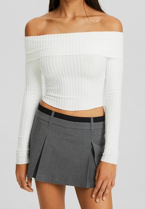 Vrouw draagt een witte off-shoulder geribbelde cropped top gecombineerd met een hooggetailleerde grijze geplooide rok met een zwarte tailleband.