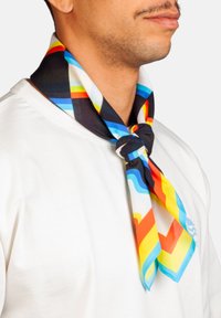 Hombre con una camisa blanca que lleva un pañuelo colorido anudado al cuello, con rayas geométricas en negro, azul, amarillo, rojo y blanco.
