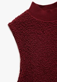 Bordeaux farvet struktureret sweater med en ribbet høj hals, med et unikt krøllemønster og ærmeløs design. Blødt strikket stof.
