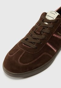 Sneakers en daim marron avec des lacets marron foncé, des accents roses et une semelle en caoutchouc texturée. Ils présentent un design rétro et un étiquetage fait à la main.