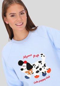 Világoskék pulóver színes hímzett kiskutya mintával. A szöveg "Mymo Pop" piros betűkkel, pöttyös és színes tappancs díszítésekkel.