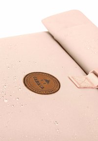 Helle rosa Rucksack mit einer strukturierten, wasserdichten Außenseite, einem runden braunen Logopatch und einem hellen Trageakzent.