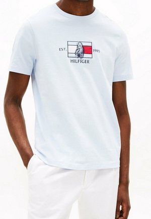 Mann trägt hellblaues Kurzarmhemd mit rotem, weißem und marineblauem Grafikmotiv und dem Text "EST. 1985 HILFIGER" auf der Brust.