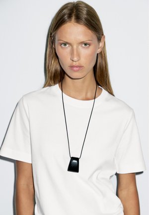 Collier pendentif noir sur un cordon noir porté sur un t-shirt blanc. Le pendentif présente un design lisse et angulaire avec une finition brillante.