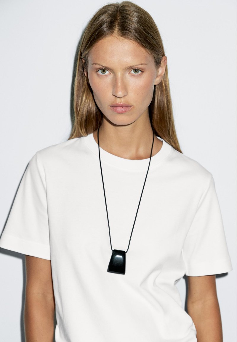 Collana con pendente nero su un cordino nero indossata sopra una maglietta bianca. Il pendente presenta un design angolare e liscio con una finitura lucida.