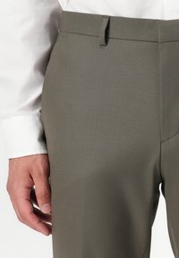 Khaki dress pants with a smooth texture, featuring a waistband with a hook and eye closure. Paired with a white shirt cuff in the background.