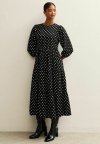 Zwart jurkje met stippen, met een getailleerde taille, gelaagde rok en lange poffende mouwen. G gedragen met hoge zwarte laarzen met een glanzende afwerking.