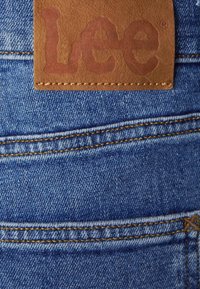 Tissu en denim bleu clair avec des coutures jaunes, comportant un patch en cuir marron embossé avec "Lee". Conception minimaliste des poches arrière.