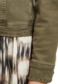 Chaqueta verde oliva con dobladillo deshilachado, con cierre de botones y textura de mezclilla, combinada con un vestido de patrón tie-dye multicolor.