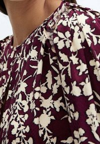 Blouse florale avec un fond prune et des motifs botaniques blancs, texture plissée et encolure ronde. Le tissu semble léger.