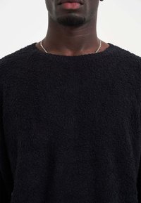 Zwarte, getextureerde trui met een losse pasvorm, ronde halslijn en lange mouwen. Bevat een zachte, donzige stof. Metalen ketting zichtbaar bij de hals.