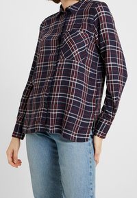 Chemise à carreaux bleu marine et rouge à manches longues, avec une poche sur la poitrine et une texture de tissu douce. Détails en lignes blanches.