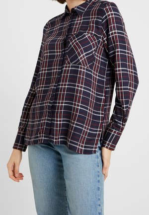 Chemise à carreaux bleu marine et rouge à manches longues, avec une poche sur la poitrine et une texture de tissu douce. Détails en lignes blanches.