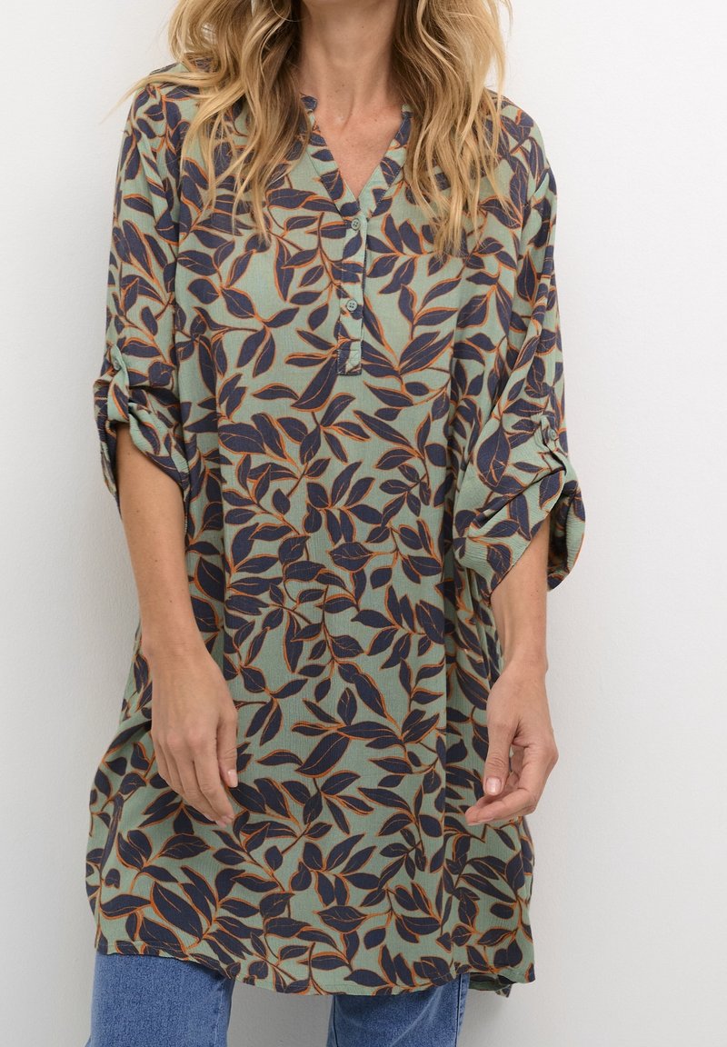 Femme portant une tunique verte ample avec des motifs de feuilles bleu foncé et orange et des manches retroussées, associée à un jean bleu.
