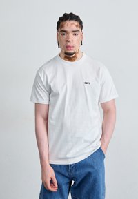 Fehér pamut póló rövid ujjakkal, bal melle környékén egy kis fekete "OBEY" logóval, kék farmer rövidnadrággal viselve.
