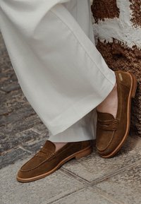 Mocassini in suede marrone con un design in stile mocassino, dotati di un'apertura cucita e una suola in gomma marrone chiaro. Indossati con pantaloni bianchi.