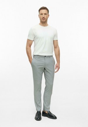 Man staat met handen in zijn zakken, draagt een wit shirt met korte mouwen, lichtgrijze broek en zwarte nette schoenen op een witte achtergrond.