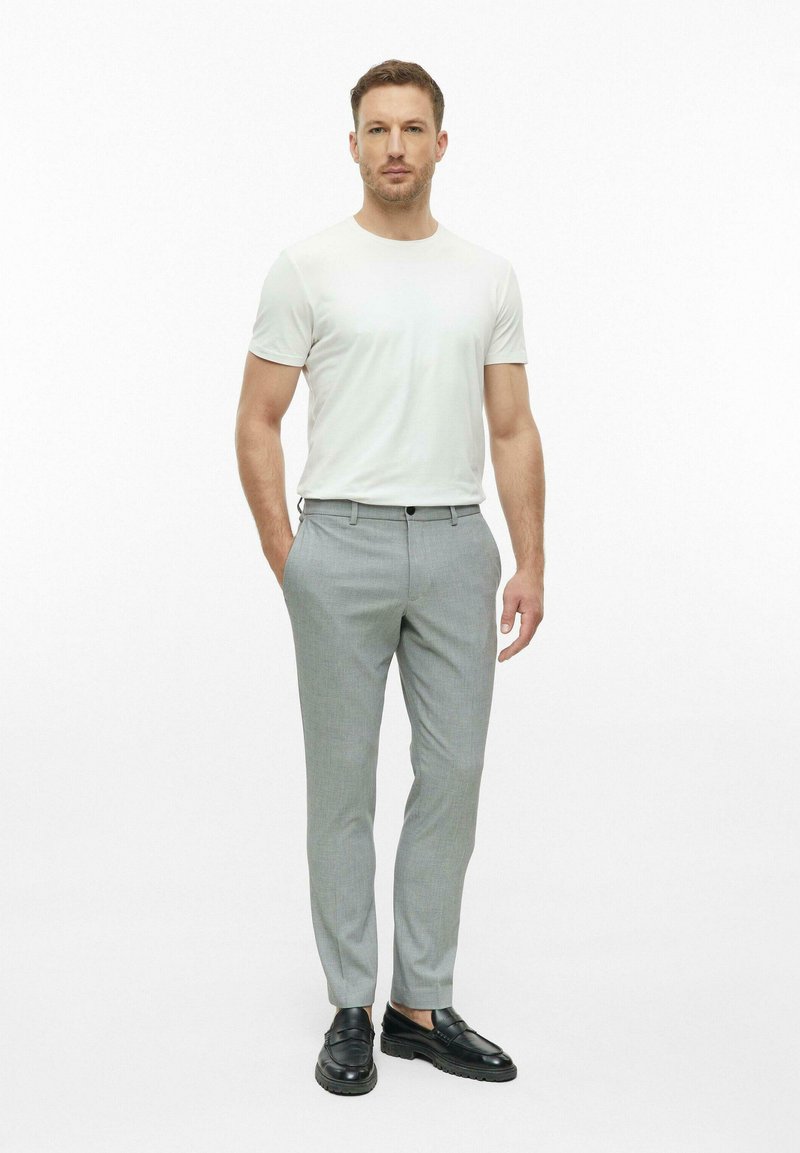 Man standing with hands in pockets wearing white short-sleeve shirt, light gray trousers, and black dress shoes on white background.