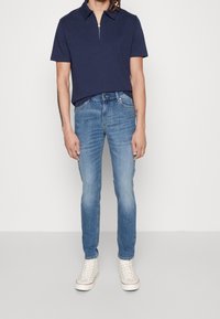 Marinblå polo med dragkedja i kragen, tillsammans med ljusblå slim-fit jeans. Modellen bär vita högskurna sneakers.