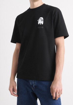 Person iført sort t-shirt med korte ærmer med lille hvidt spøgelsesmotiv og teksten "HUGO" på brystet, kombineret med blå jeans.