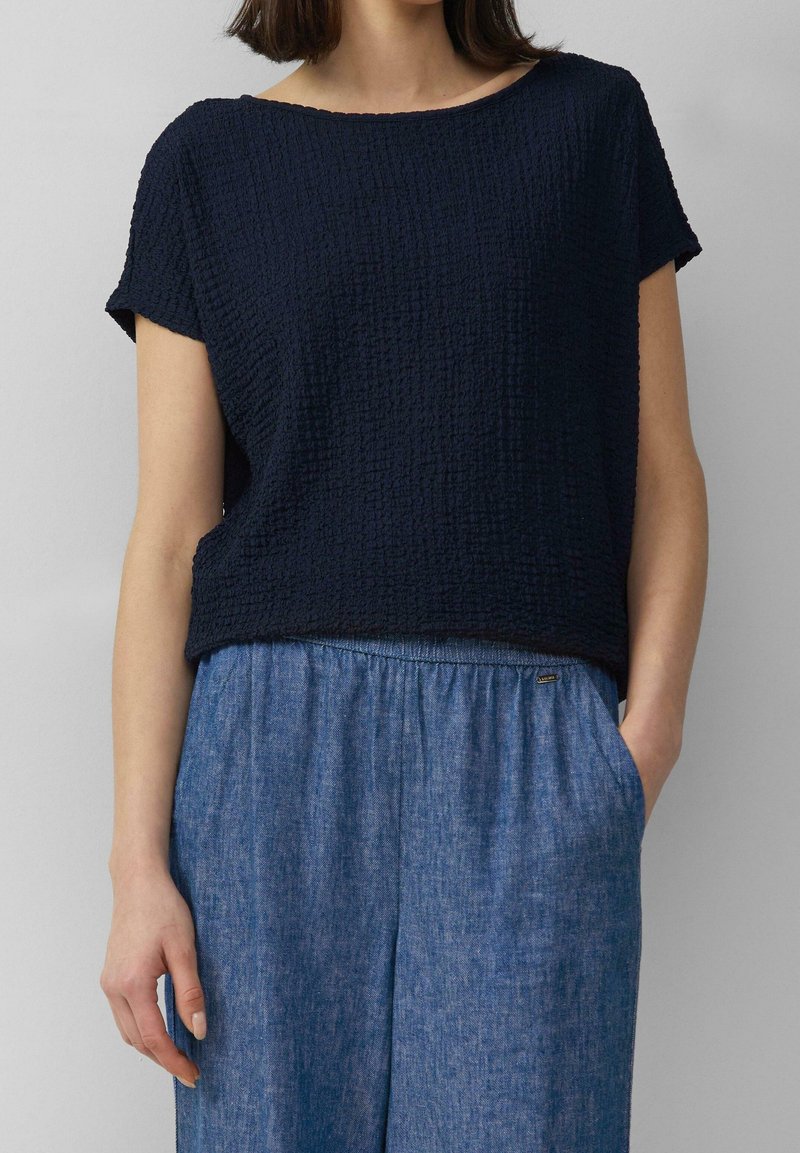 Femme portant un haut texturé bleu marine à manches courtes et un pantalon bleu ample, une main dans la poche, sur fond gris.