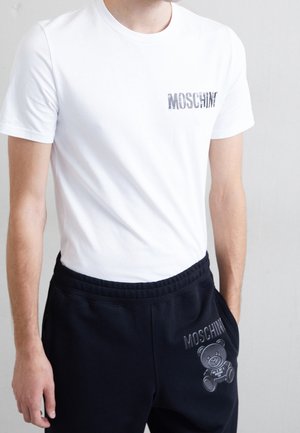 Homme portant un t-shirt blanc Moschino et un pantalon de jogging noir avec le logo Moschino et un motif d'ours en peluche, debout devant un fond uni.