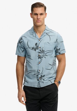 Mężczyzna noszący jasnoniebieską, krótkorękawową koszulę z ciemnymi kwiatowymi wzorami i czarne spodnie, stojący z jedną ręką w kieszeni.