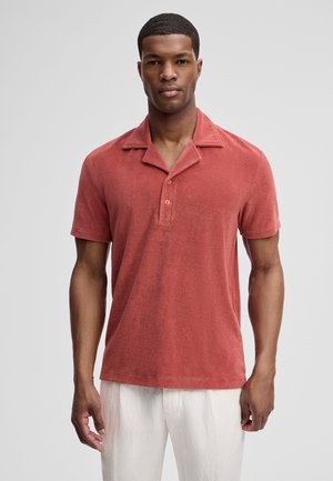 Mann trägt ein kurzärmeliges, rostfarbenes Polohemd mit zwei Knöpfen und weiße Hose, steht vor einem schlichten grauen Hintergrund.