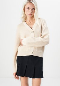 Beige geribde cardigan met een V-hals, sluiting met knop aan de voorkant en twee zakken. Draagt over een zwarte geplisseerde rok, met lange mouwen.