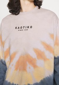 Dlouhý rukáv tričko s batikovaným vzorem v béžových, oranžových a modrých odstínech s textem "KAOTIKO SINCE 1999" raženým na hrudi. Měkká bavlněná tkanina.