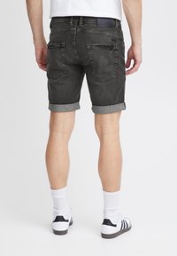 Mand iført opkrøllede mørk denim shorts, hvide sokker og hvide sneakers set fra ryggen mod en ensfarvet baggrund.