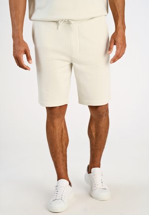 Cremefarbene Shorts aus weichem Material, mit elastischem Bund und Kordelzug sowie umgeschlagenen Bündchen. Kombiniert mit weißen Turnschuhen.