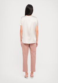 Donna con lunghi capelli scuri che indossa un top bianco a maniche corte e morbido, pantaloni su misura color malva e mule bianche con tacco a rocchetto, in piedi davanti a uno sfondo semplice.