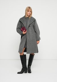 Grauer Wollmantel mit großem Kragen, oversized Passform und einer Knopfverschluss. Das Model trägt ein graues Kleid und schwarze knielange Stiefel.