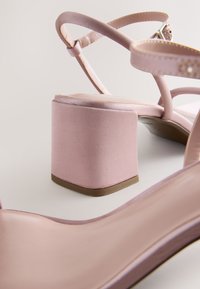 Sandali con tacco alto in satin rosa con un largo tacco a blocco, cinturini incrociati e finitura liscia e lucida. Rifiniti con sottili dettagli metallici.