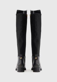 Une paire de bottes noires montantes avec des talons bas, des tirettes dorées et un logo doré MK à l'arrière de la zone en cuir du talon.