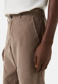 Brune bukser i manchestermateriale med elastisk taljebånd, der har en sort knap, sidelommer og en tekstureret stoffinish.