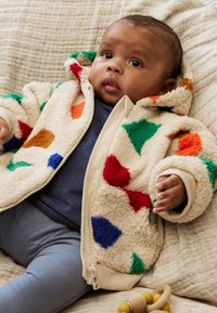 Veste bébé zippée en polaire, crème avec des formes géométriques colorées en rouge, vert, orange et bleu ; texture douce et capuche.